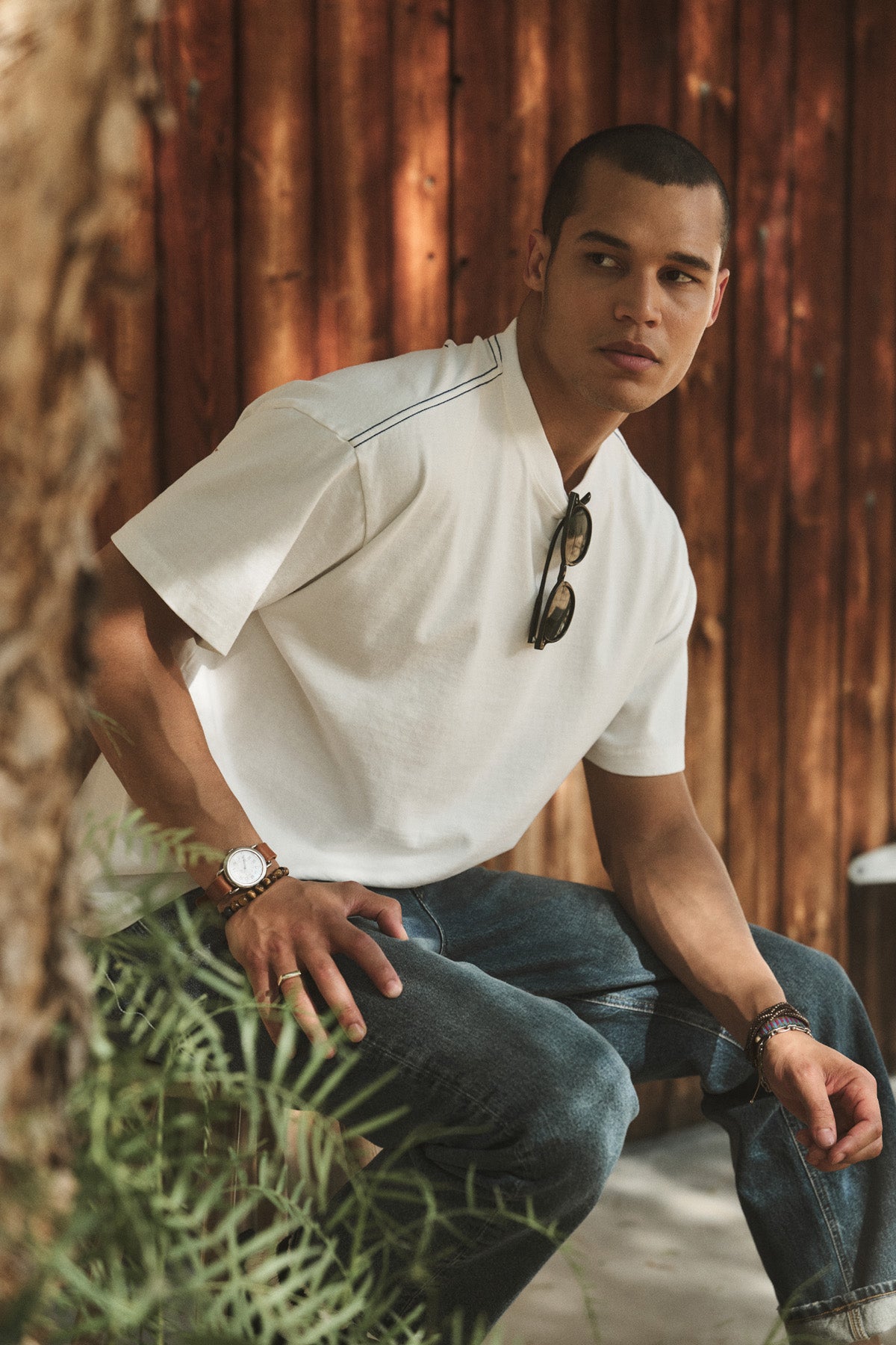 A man sits outdoors by a tree in the boxy, relaxed fit white ISAAC TEE from Velvet by Graham & Spencer and jeans, with sunglasses hanging from his shirt and a wooden wall behind him.-41105029660865