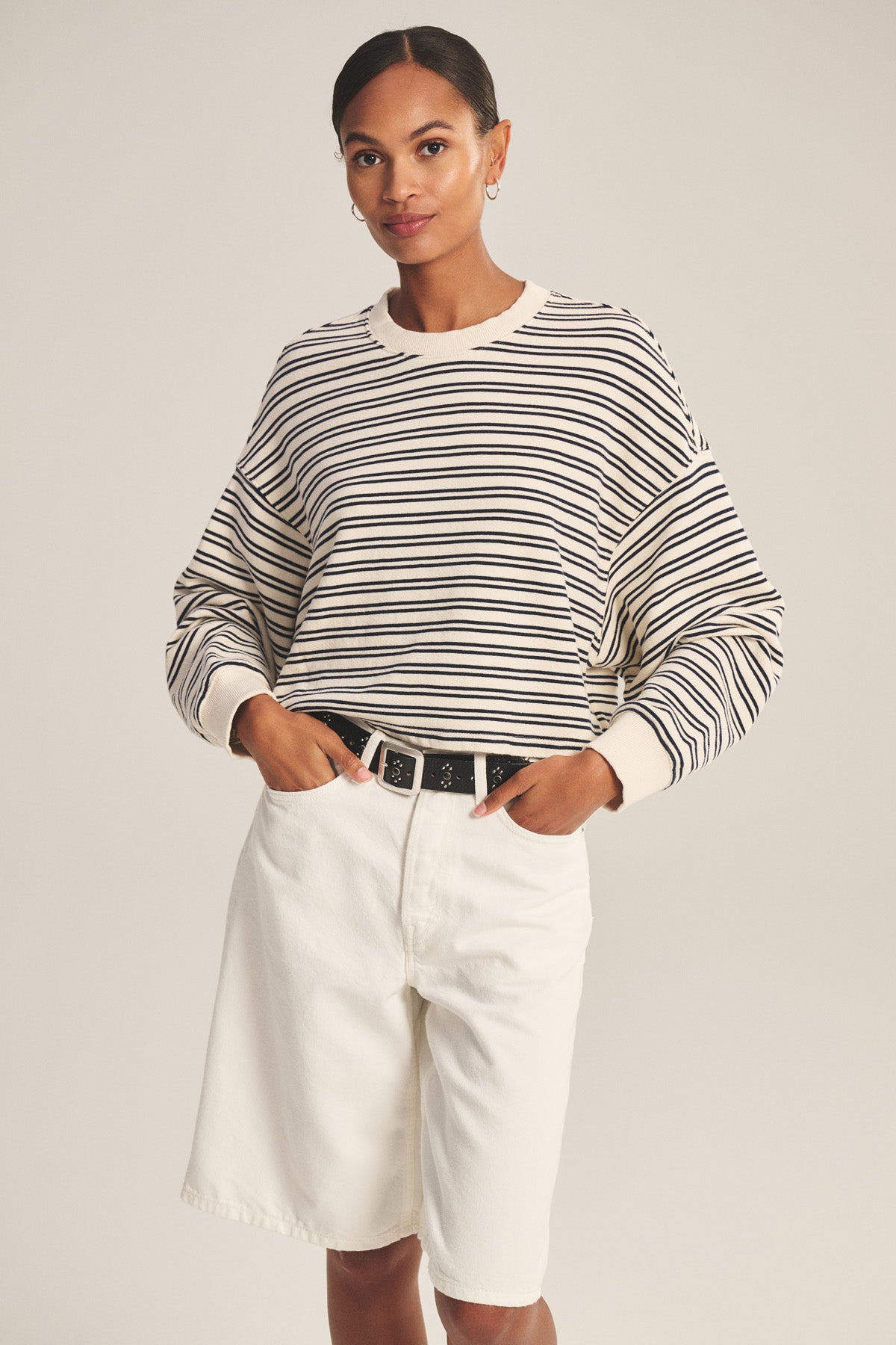 A woman poses against a plain background wearing the ISABELLE PULLOVER by Velvet by Graham & Spencer, styled with high-waisted white shorts and a black belt for a chic, casual look.