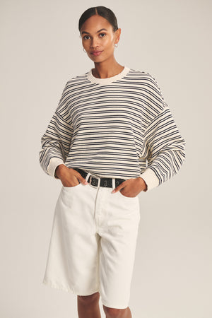 A woman poses against a plain background wearing the ISABELLE PULLOVER by Velvet by Graham & Spencer, styled with high-waisted white shorts and a black belt for a chic, casual look.