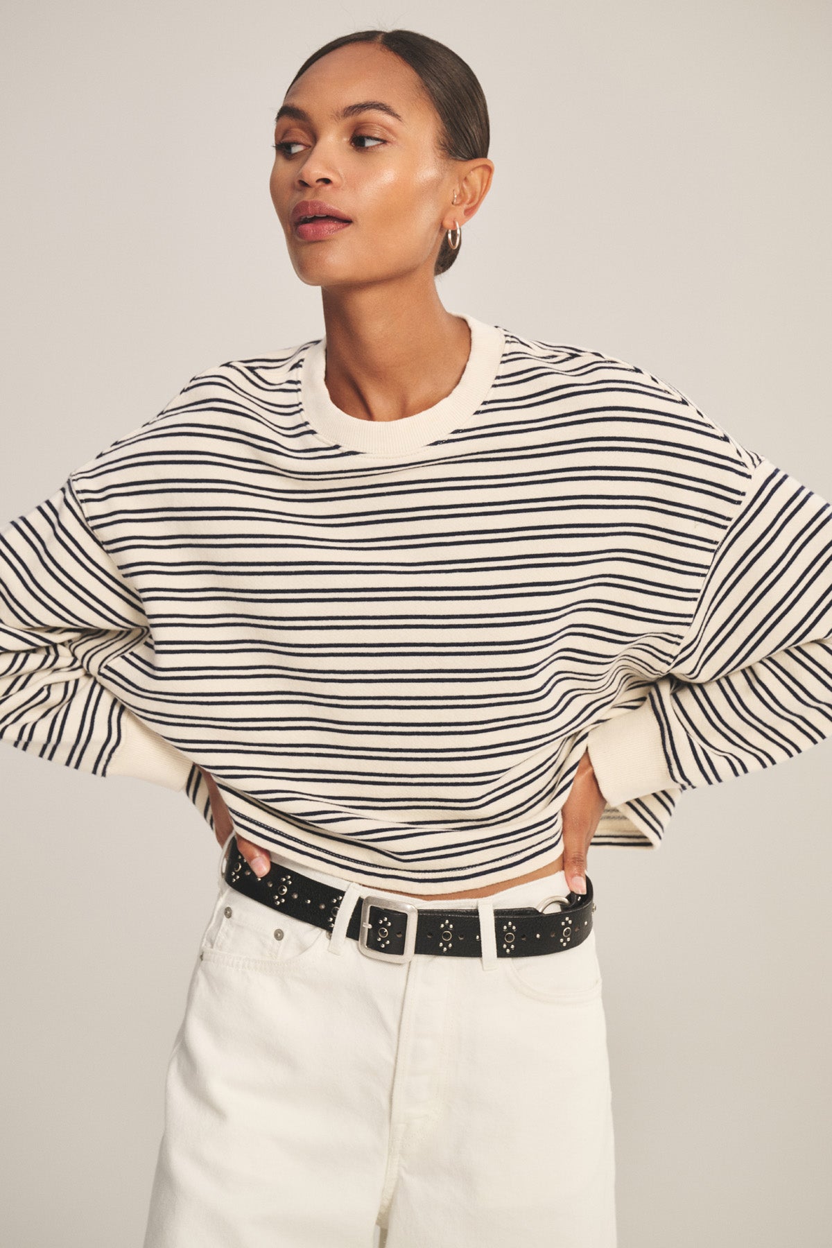 A model wears the ISABELLE PULLOVER by Velvet by Graham & Spencer—a black-and-white stripe terry cropped top—paired with white pants and a black belt, standing hands on hips to showcase relaxed, chic style against a neutral background.-41088941359297