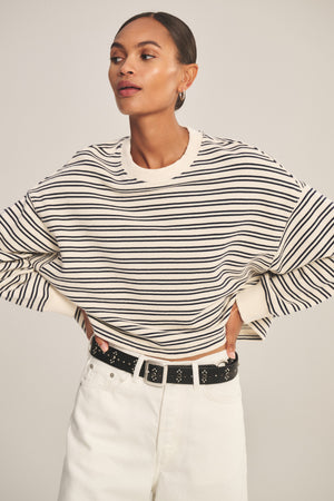 A model wears the ISABELLE PULLOVER by Velvet by Graham & Spencer—a black-and-white stripe terry cropped top—paired with white pants and a black belt, standing hands on hips to showcase relaxed, chic style against a neutral background.