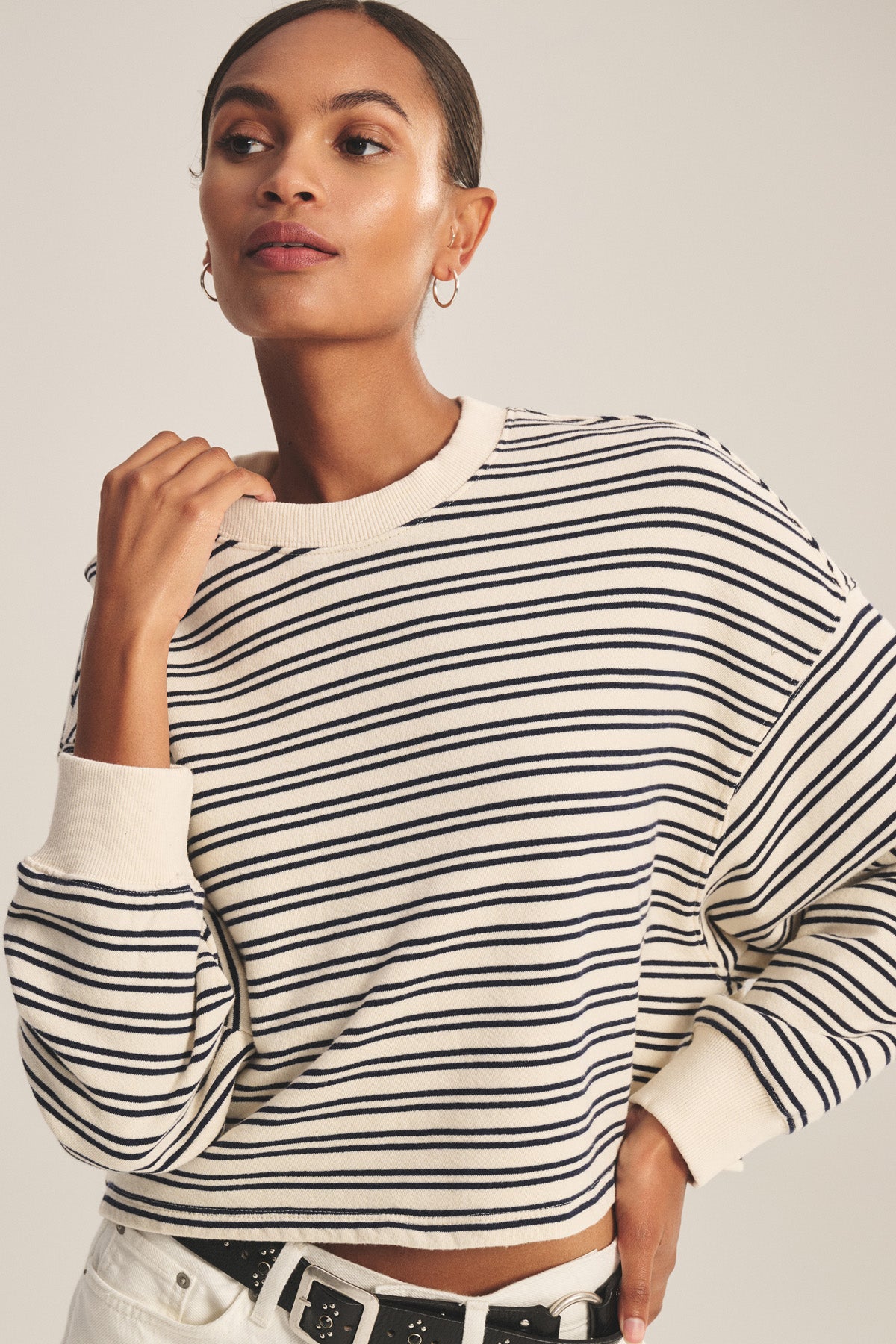 Wearing the ISABELLE PULLOVER by Velvet by Graham & Spencer—a cream, black-striped terry long-sleeve cropped top—paired with white pants and a black belt against a neutral background. Perfect for casual style.-41088941326529