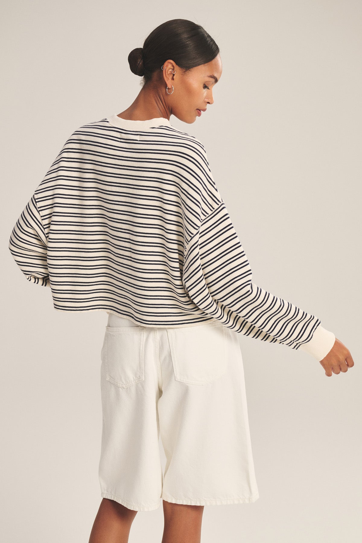 A woman stands turned away, wearing the Velvet by Graham & Spencer ISABELLE PULLOVER and white shorts, her hair pulled back in a neat bun for a casual look.-41088941293761