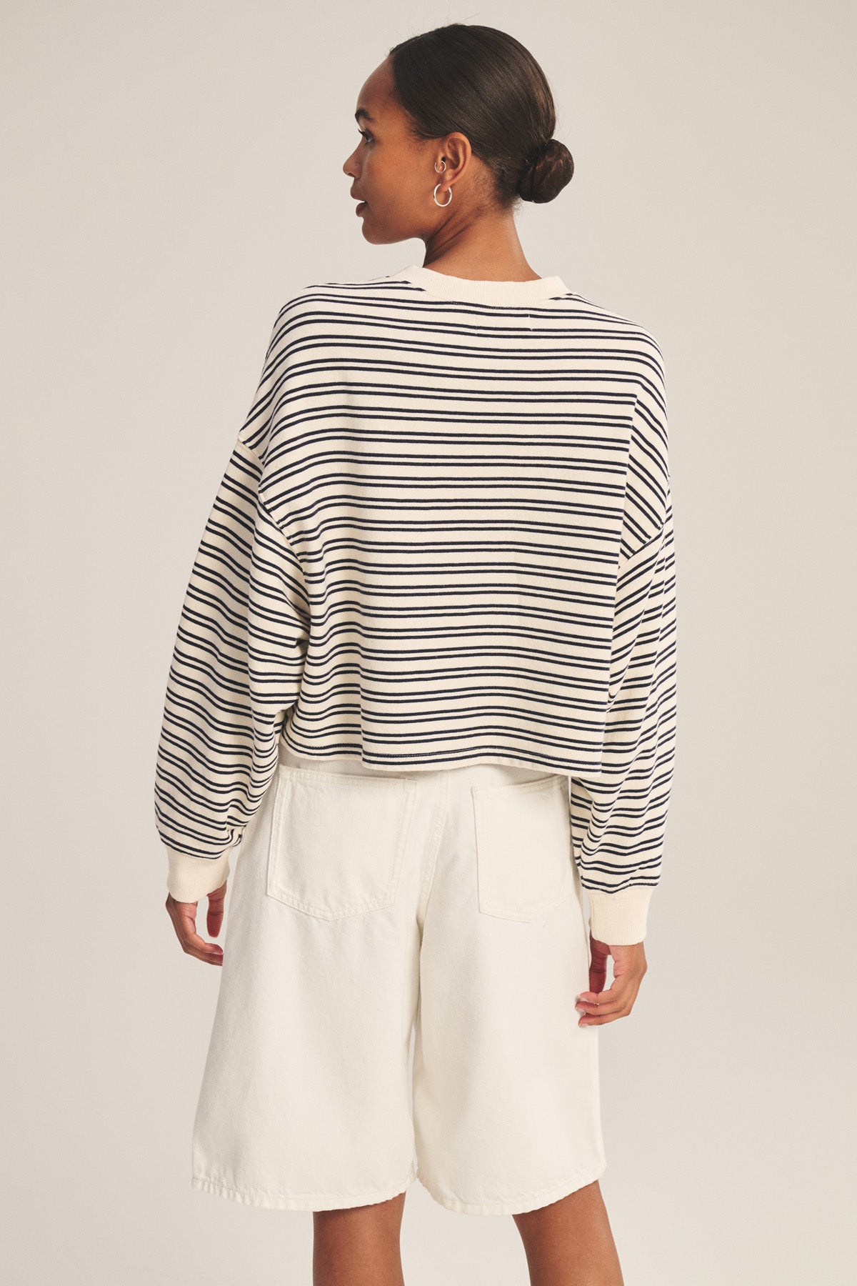 Person stands with back to camera in Velvet by Graham & Spencer’s ISABELLE PULLOVER, a black and white striped terry long-sleeve top, paired with white knee-length shorts against a plain background for a chic casual look.-41088941260993