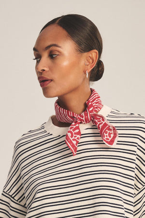 A woman with a sleek bun wears Velvet by Graham & Spencer’s LISETTE SCARF in red patterns and a black and white striped shirt, looking to the side against a plain background.