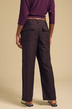 A person stands against a beige background wearing the JACLYN PANT by Velvet by Graham & Spencer—wide-leg, dark brown pants crafted from brushed cotton sateen and featuring large back pockets—paired with a purple long-sleeve top and flat shoes. The cropped silhouette of the ensemble lends a modern touch to the look.