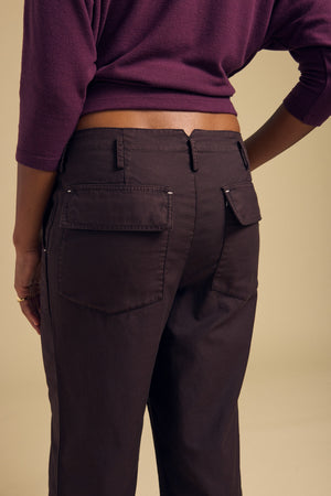 A person is shown from the back against a beige background, wearing the JACLYN PANT by Velvet by Graham & Spencer in dark brown brushed cotton sateen with a wide leg silhouette, paired with a purple top and their hands by their sides.