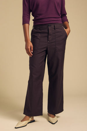 A person stands against a beige background wearing the Velvet by Graham & Spencer JACLYN PANT, a dark, wide-leg style in a cropped silhouette crafted from soft brushed cotton sateen with front pockets, paired with a purple top and cream-colored shoes.
