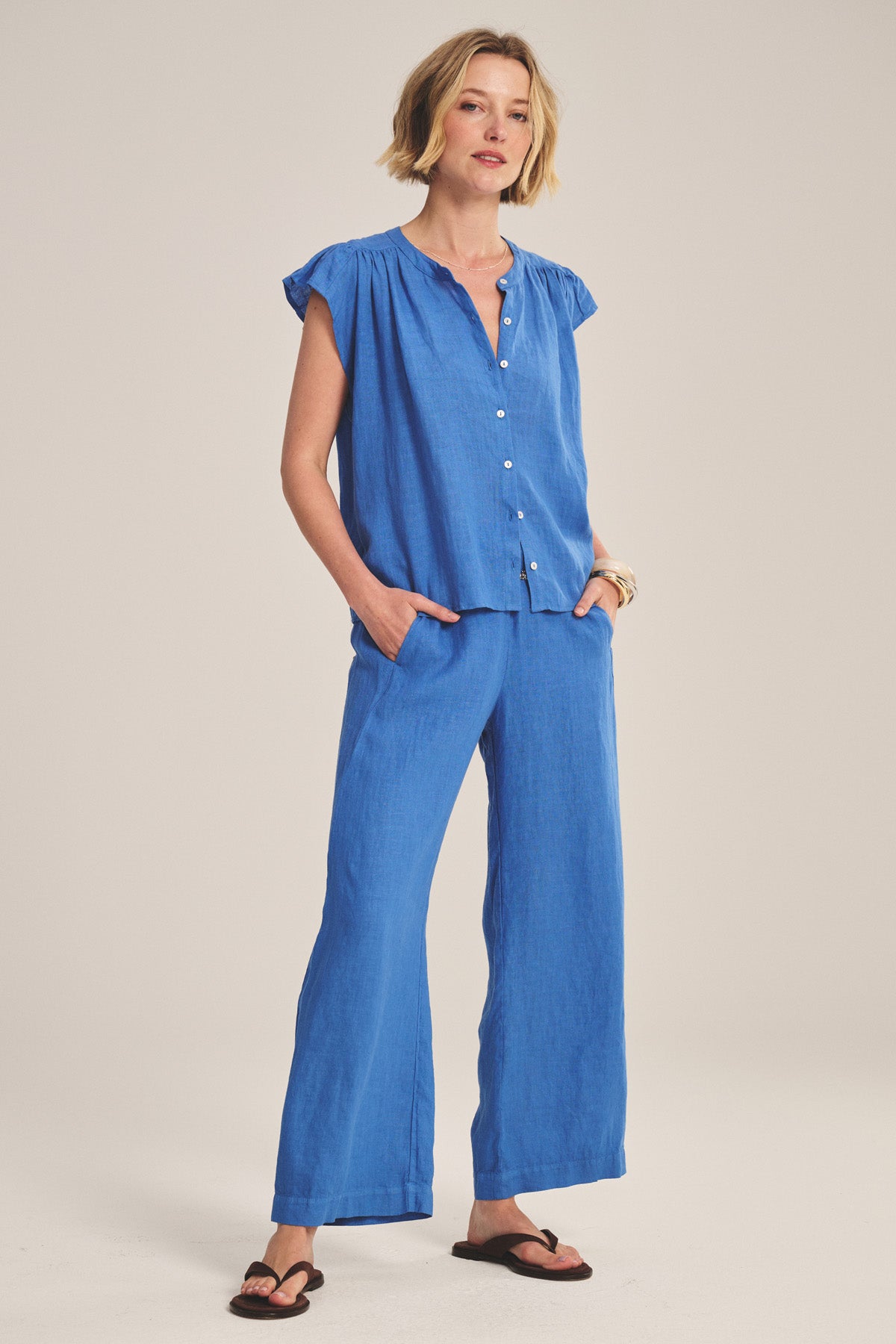 A woman wears the JANEY LINEN TOP by Velvet by Graham & Spencer, featuring a button-front closure and breathable fabric, styled with matching wide-leg pants and black sandals against a neutral background.