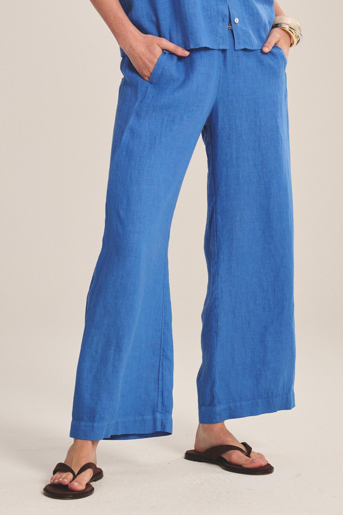 Wearing the Velvet by Graham & Spencer LOLA LINEN PANT with an elastic waist, a matching blue top, black flip-flops, and a bracelet, this person stands with one hand in their pocket against a plain background.-41088973865153