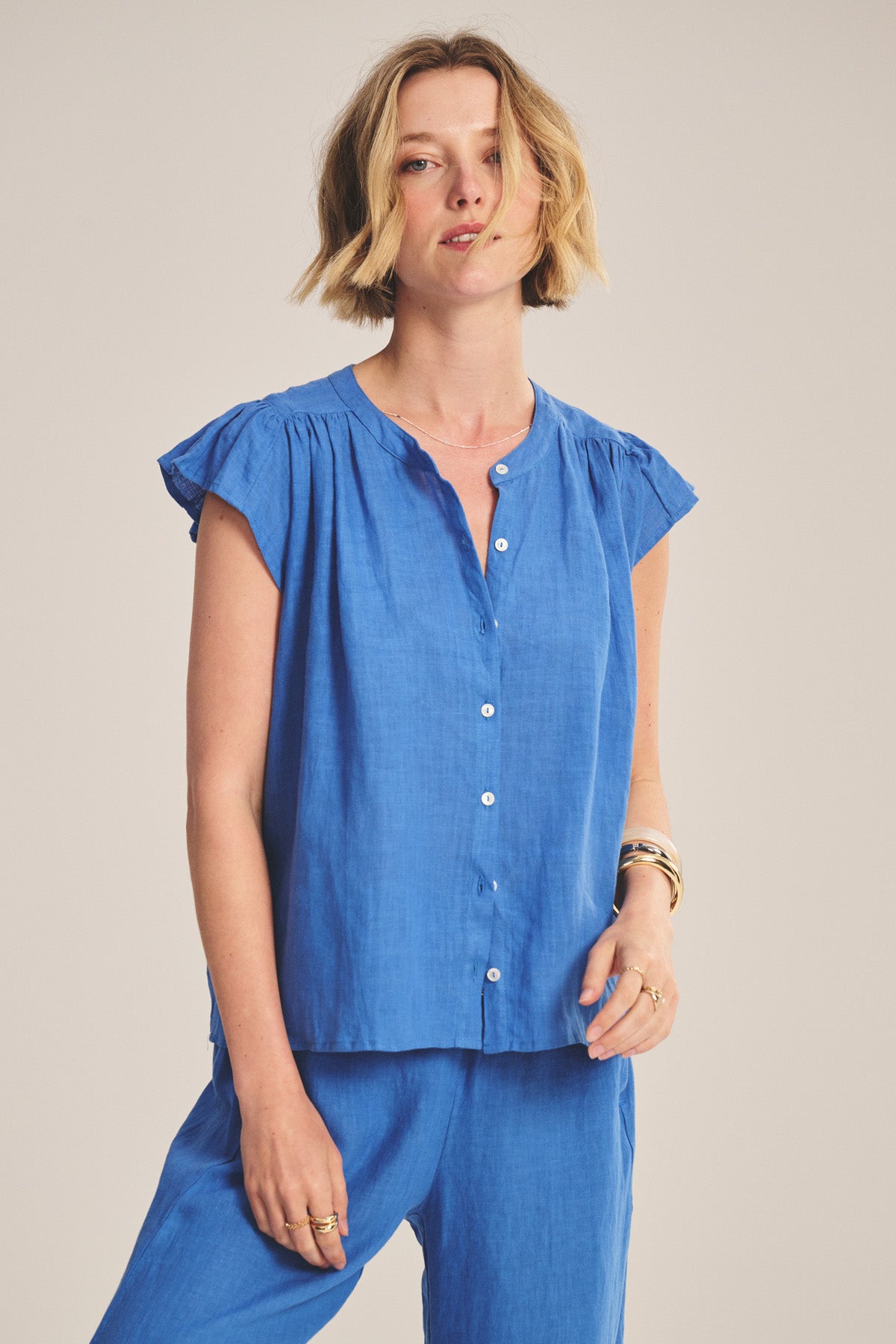 A woman wears the JANEY LINEN TOP by Velvet by Graham & Spencer, featuring flutter sleeves and a button-front closure, paired with matching blue pants against a plain background.