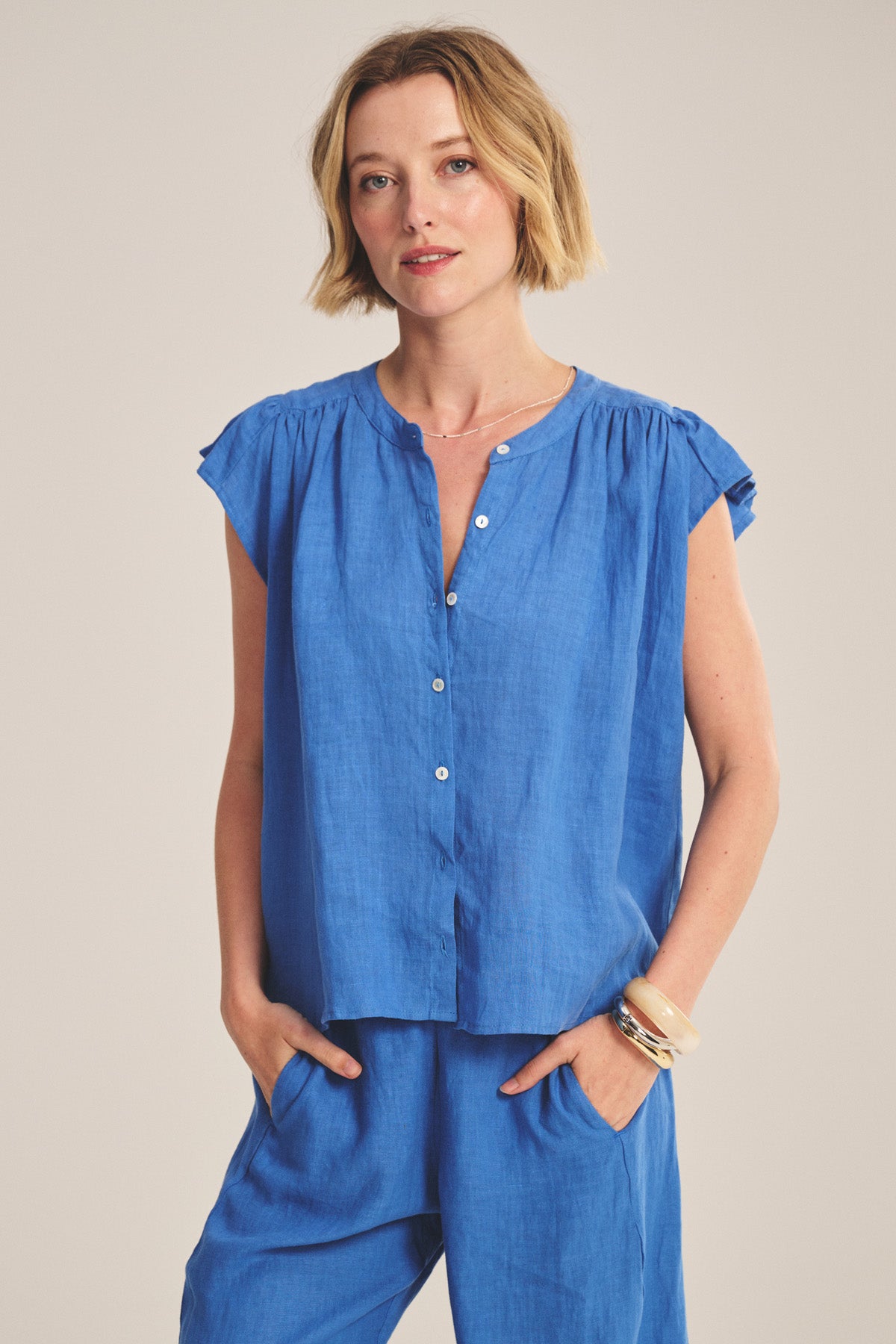 A woman wears the JANEY LINEN TOP by Velvet by Graham & Spencer—a short-sleeved, button-front blue blouse paired with matching pants—as she stands with hands in her pockets against a plain background.