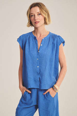 A woman wears the JANEY LINEN TOP by Velvet by Graham & Spencer—a short-sleeved, button-front blue blouse paired with matching pants—as she stands with hands in her pockets against a plain background.