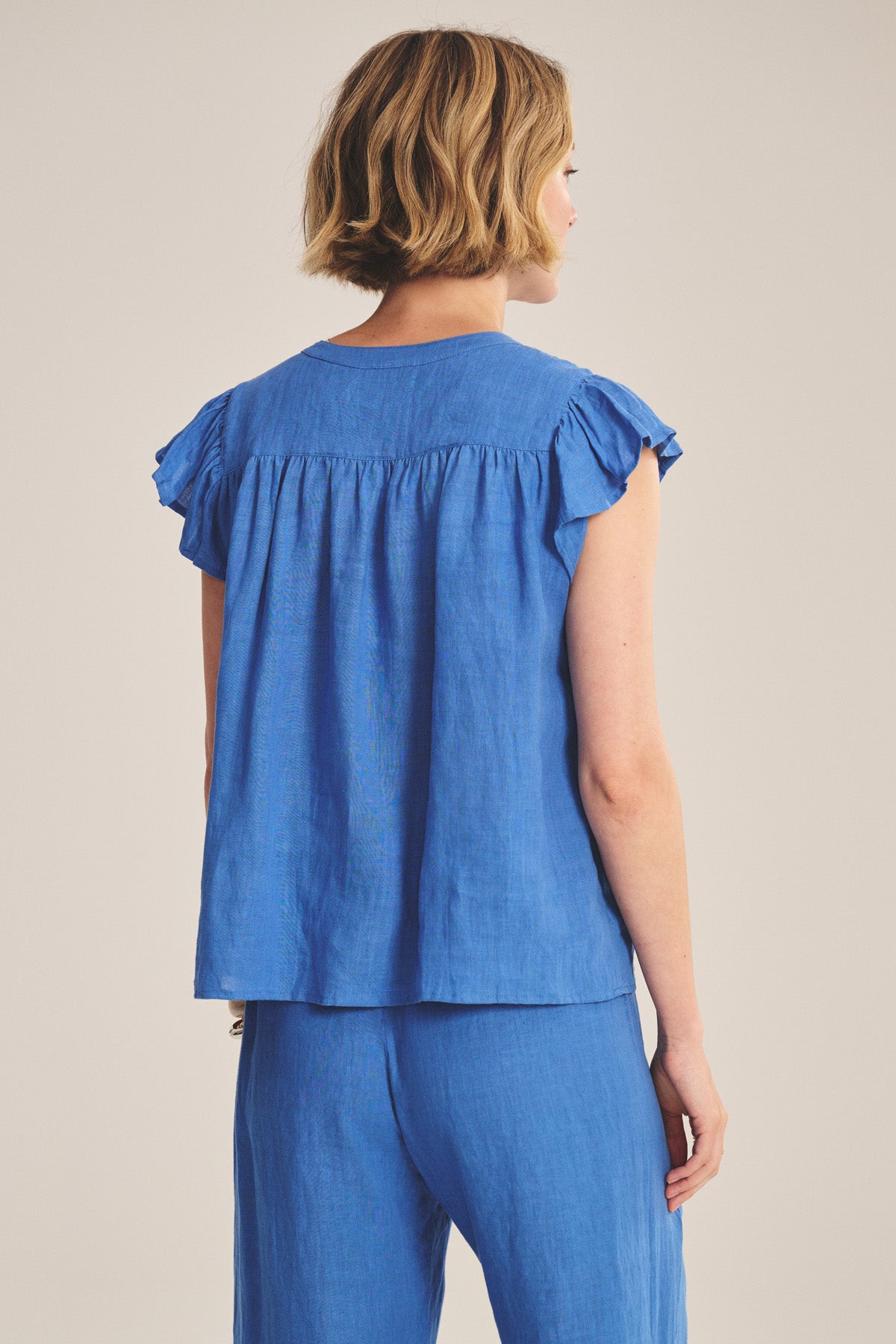 A person with short blonde hair is shown from the back, wearing the JANEY LINEN TOP by Velvet by Graham & Spencer—a sleeveless blue top with ruffled shoulders and button-front closure—paired with matching blue pants.