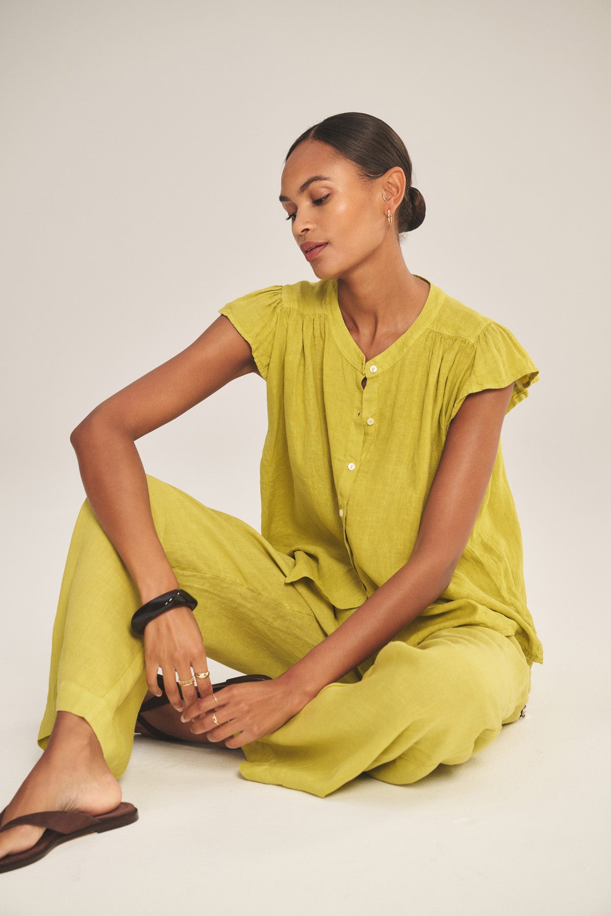 A woman sits on the floor wearing the JANEY LINEN TOP by Velvet by Graham & Spencer—a loose-fitting yellow button-front, breathable linen top—paired with brown sandals and a black bracelet.