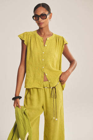 A woman wears the Velvet by Graham & Spencer JANEY LINEN TOP in light green with a button-front, styled with matching wide-leg pants and a coordinating jacket, accessorized with oversized sunglasses and a black bracelet.