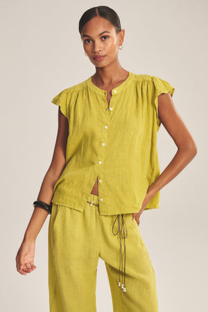 A woman wears the JANEY LINEN TOP by Velvet by Graham & Spencer, featuring a breathable fabric and button-front closure, paired with matching wide-leg pants. She stands with one hand in her pocket against a neutral background.