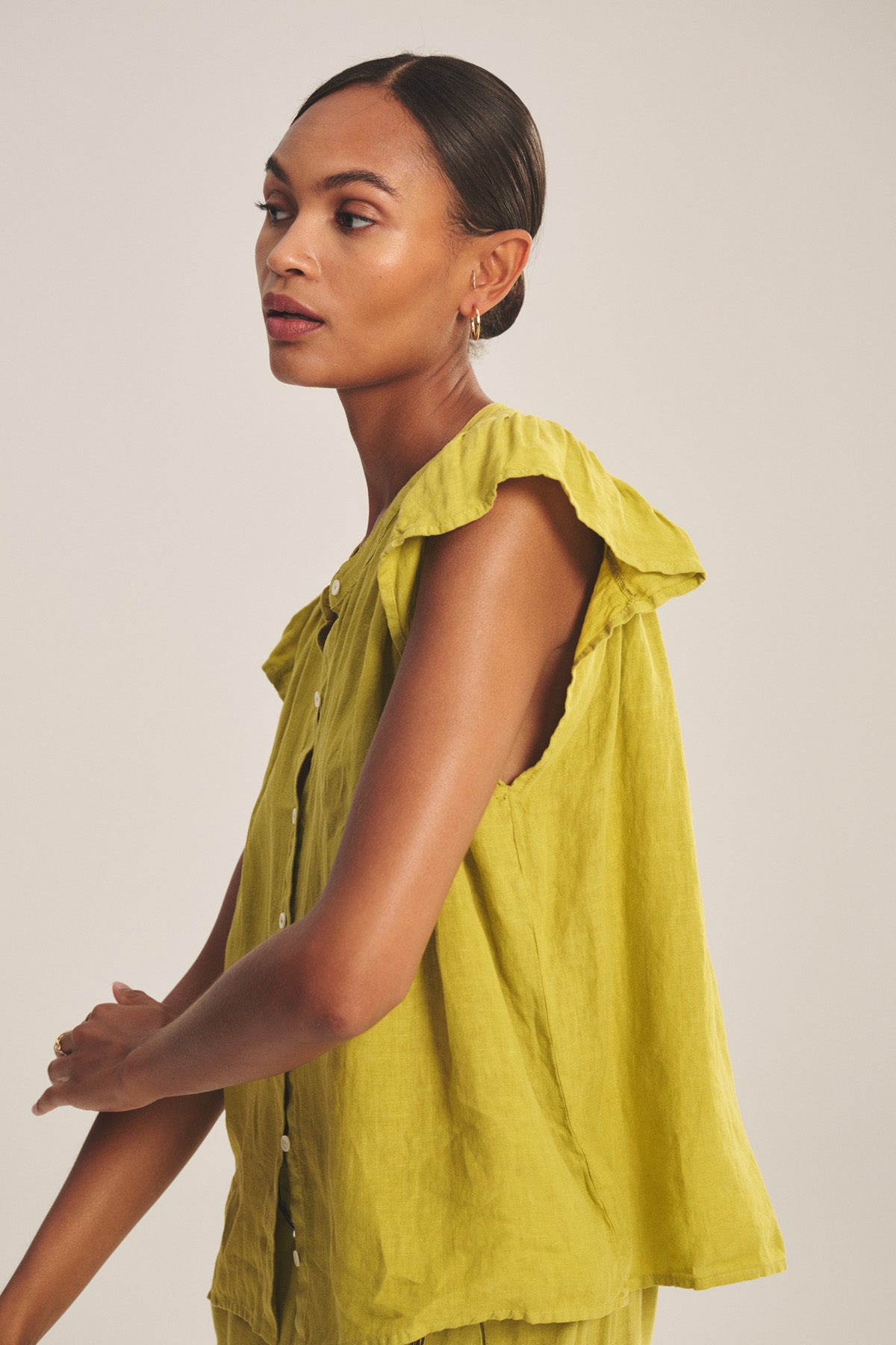 A woman stands against a plain background wearing the JANEY LINEN TOP by Velvet by Graham & Spencer, a sleeveless, loose-fitting lime green linen shirt with ruffled shoulders.