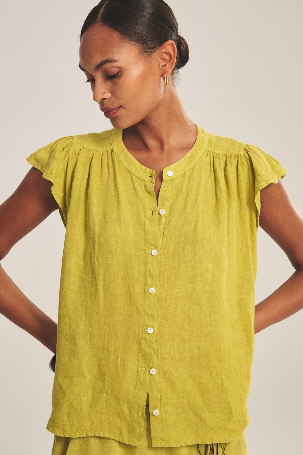 A woman wears the JANEY LINEN TOP by Velvet by Graham & Spencer, a light yellow, short-sleeved blouse with a button-front closure, standing with her arms slightly bent and gazing downward.