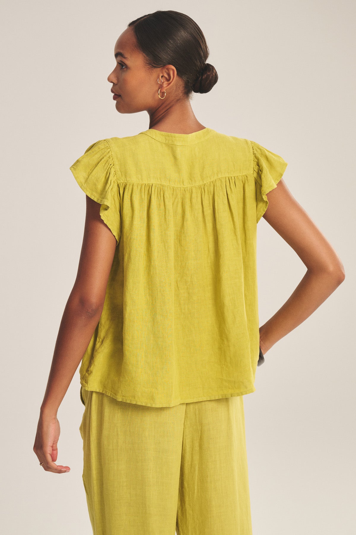 A woman stands with her back to the camera, wearing the JANEY LINEN TOP by Velvet by Graham & Spencer, a loose-fitting, breathable linen shirt with short sleeves, paired with matching yellow pants.