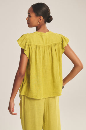 A woman stands with her back to the camera, wearing the JANEY LINEN TOP by Velvet by Graham & Spencer, a loose-fitting, breathable linen shirt with short sleeves, paired with matching yellow pants.