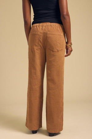A person is seen from the back wearing the JAYDA CORDUROY PANT by Velvet by Graham & Spencer—tan, wide-leg cotton corduroy pants with a drawstring elastic waist—paired with a black sleeveless top, against a plain beige background.