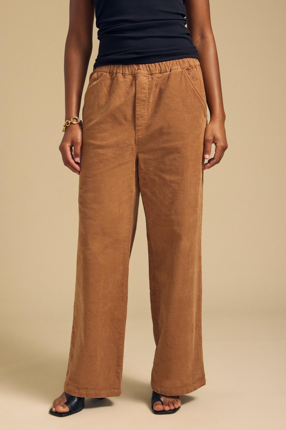 A person is seen wearing the JAYDA CORDUROY PANT by Velvet by Graham & Spencer—loose-fitting brown cotton corduroy pants with a wide-leg silhouette and a drawstring elastic waist—styled with a black sleeveless top, black open-toe heels, and a gold bracelet, standing against a plain beige background.-41964559401153