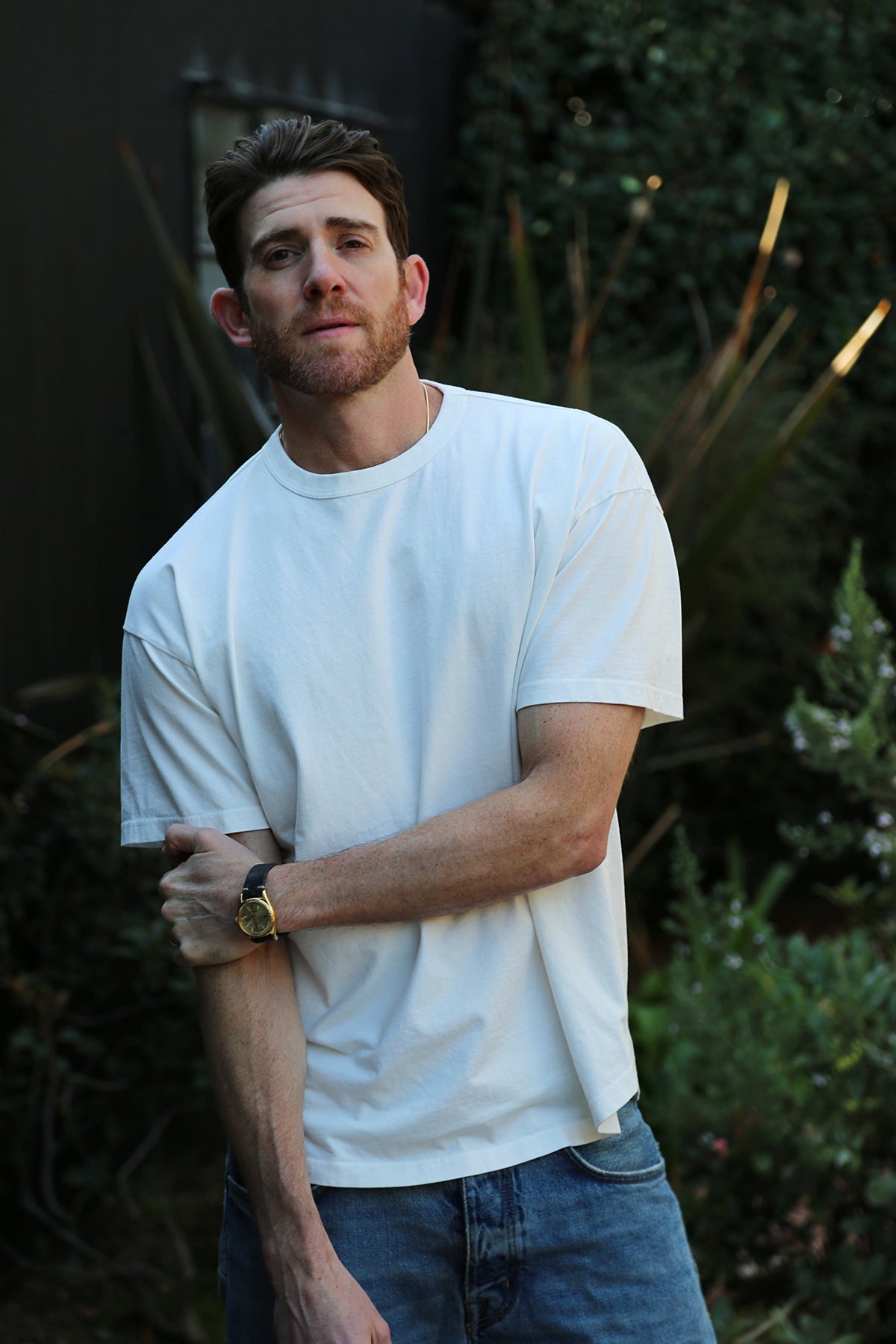   A man wearing the BRYAN TEE by Velvet by Graham & Spencer, paired with jeans, stands outdoors surrounded by greenery. 