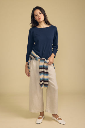 A woman stands against a beige background wearing the Velvet by Graham & Spencer GABI TEE—a navy, long-sleeve cotton jersey top with a boat neckline—paired with beige wide-leg pants, white flats, and a striped sweater tied around her waist.