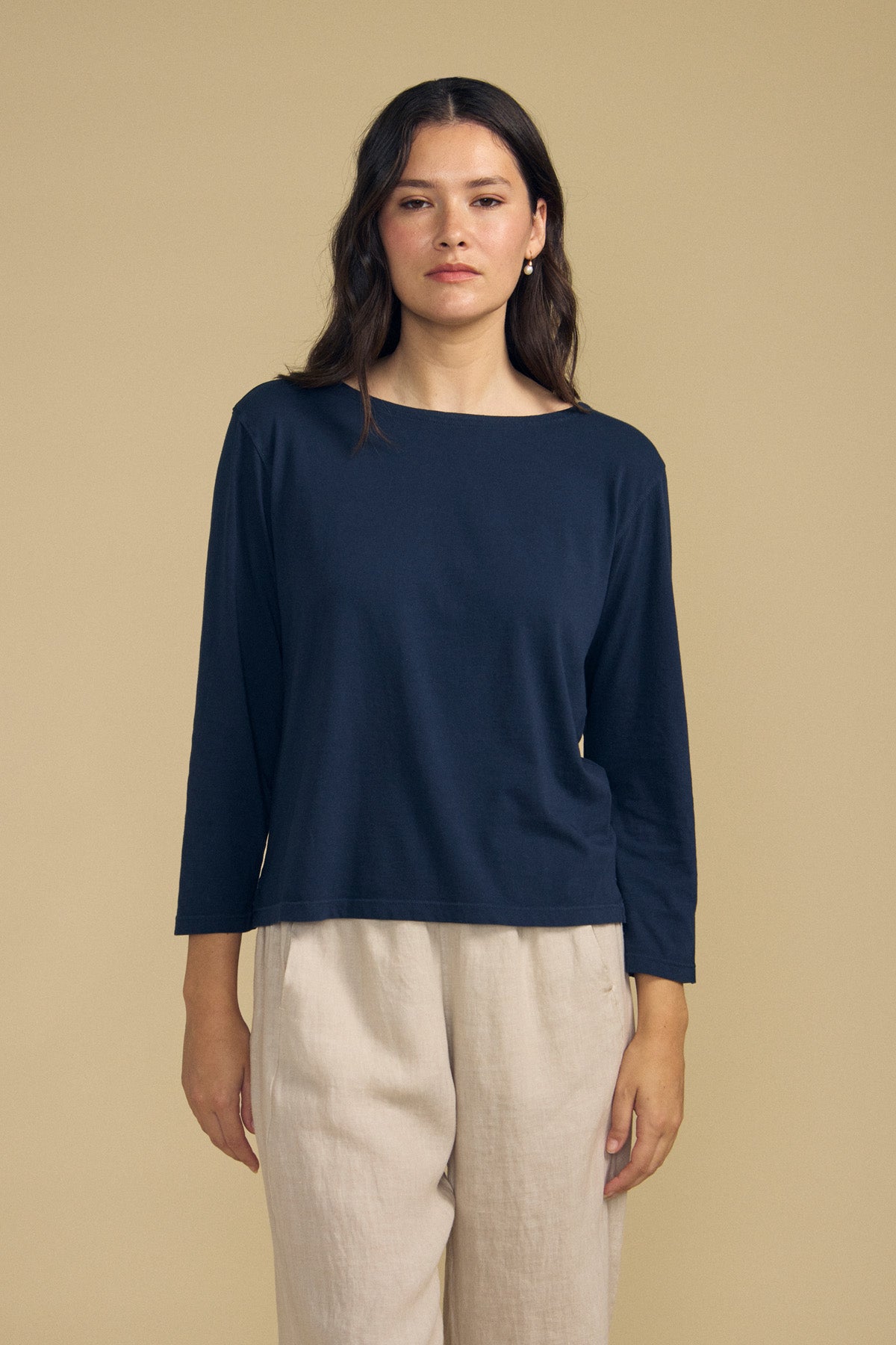 A person stands against a beige background wearing the Velvet by Graham & Spencer GABI TEE, a navy cotton jersey featuring a boat neckline, paired with light beige pants and looking straight at the camera.