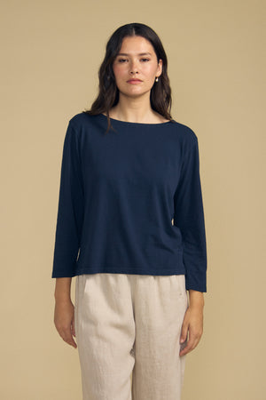A person stands against a beige background wearing the Velvet by Graham & Spencer GABI TEE, a navy cotton jersey featuring a boat neckline, paired with light beige pants and looking straight at the camera.