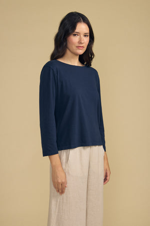 A woman stands against a beige background, wearing the GABI TEE by Velvet by Graham & Spencer—a navy cotton jersey top featuring a boat neckline and cropped length—paired with light beige wide-leg pants. She gazes slightly to the side with a neutral expression.