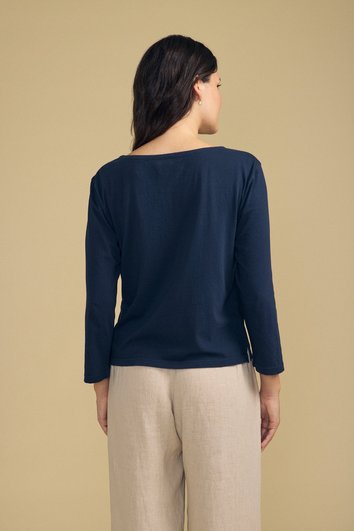 A woman stands facing away against a plain beige background, wearing the GABI TEE by Velvet by Graham & Spencer—a navy cotton jersey top with a boat neckline—paired with light beige pants.