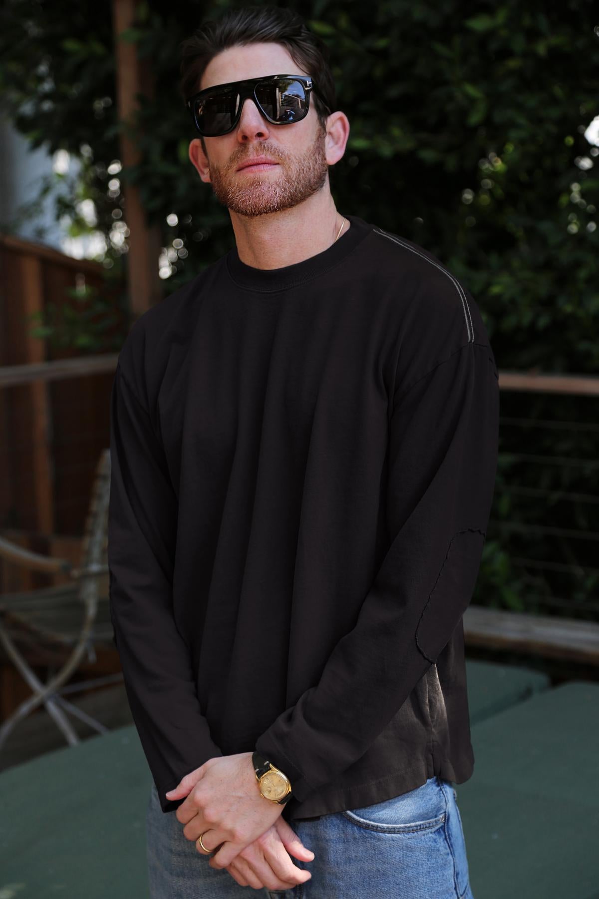 Wearing sunglasses, a SLATER TEE by Velvet by Graham & Spencer in a relaxed fit black long-sleeve made of luxurious Pima cotton jersey, and jeans, a man stands outdoors amidst greenery and a wooden railing.-38325909258433