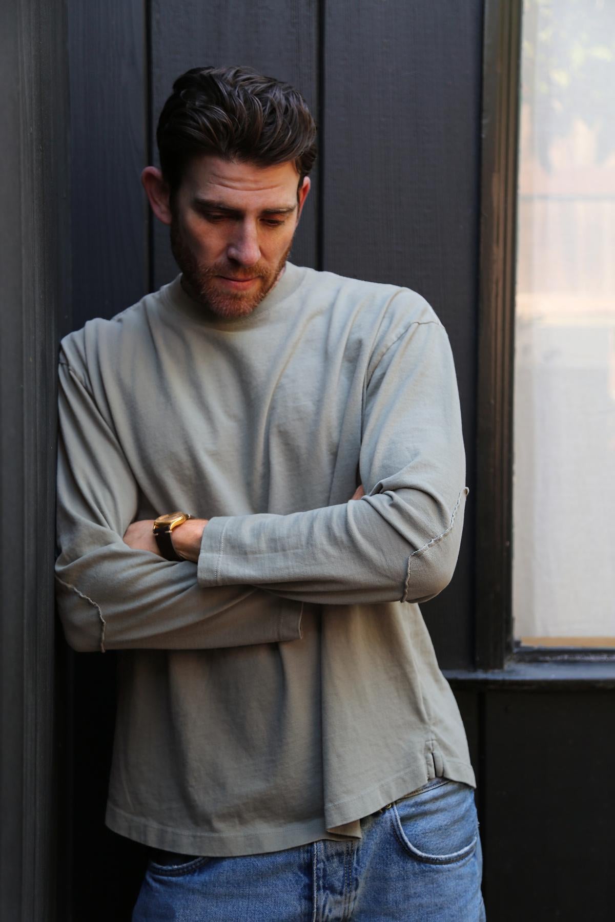   A person with dark hair and a beard leans against a black wall, looking down with arms crossed. They’re wearing the SLATER TEE from Velvet by Graham & Spencer, a light gray, ribbed crew neckline shirt made from structured pima cotton, paired with blue jeans—a versatile wardrobe addition for any occasion. 