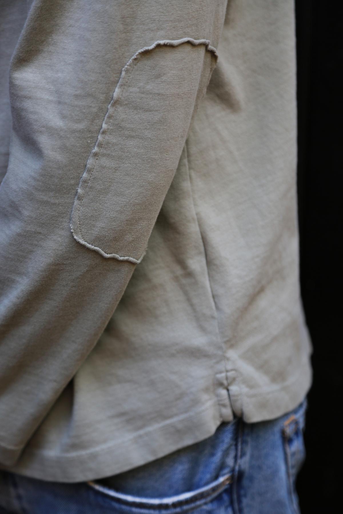   Close-up of a person wearing the SLATER TEE by Velvet by Graham & Spencer, a beige long-sleeve shirt crafted from structured pima cotton with visible stitching, paired with blue jeans. The focus is on the elbow area of the shirt, highlighting its relaxed fit as a versatile addition to any wardrobe. 