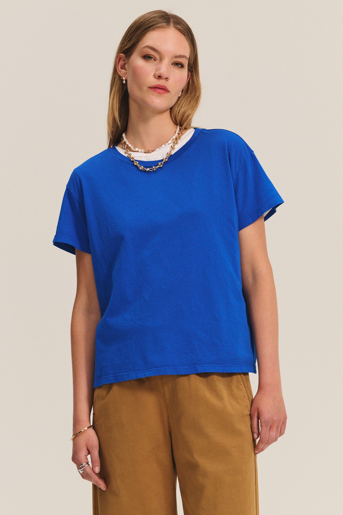 A woman in a royal blue short-sleeve tee and Velvet by Graham & Spencer’s NAYA PANT, paired with layered necklaces, stands against a plain light background.