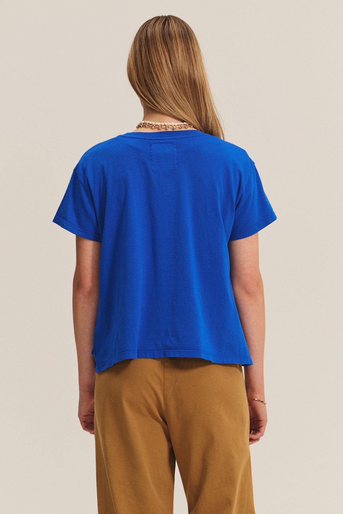 Person with long, light brown hair wearing a blue t-shirt and Velvet by Graham & Spencer NAYA PANT in tan stretch cotton twill, viewed from behind against a neutral background.