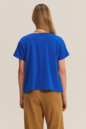 Person with long, light brown hair wearing a blue t-shirt and Velvet by Graham & Spencer NAYA PANT in tan stretch cotton twill, viewed from behind against a neutral background.