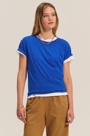 A woman wearing the NAYA PANT by Velvet by Graham & Spencer—tan wide-leg pants with an elastic waistband—paired with a blue t-shirt over a white shirt and a gold chain necklace, stands against a plain light background.