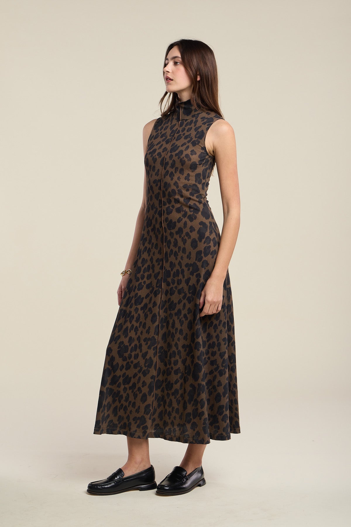 A woman stands wearing the KELESTINE DRESS by Velvet by Graham & Spencer—a sleeveless, high-neck, ankle-length dress featuring a brown and black leopard print—paired with black loafers.