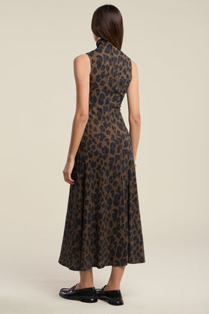 A woman stands facing away, wearing the KELESTINE DRESS by Velvet by Graham & Spencer—a sleeveless, high-neck, ankle-length leopard-print dress with an a-line skirt—paired with black shoes.