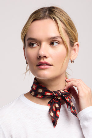 A woman with light hair wears a white top and the JEWEL SCARF by Velvet by Graham & Spencer, styled around her neck as a chic accessory, looking to the side with a neutral expression.