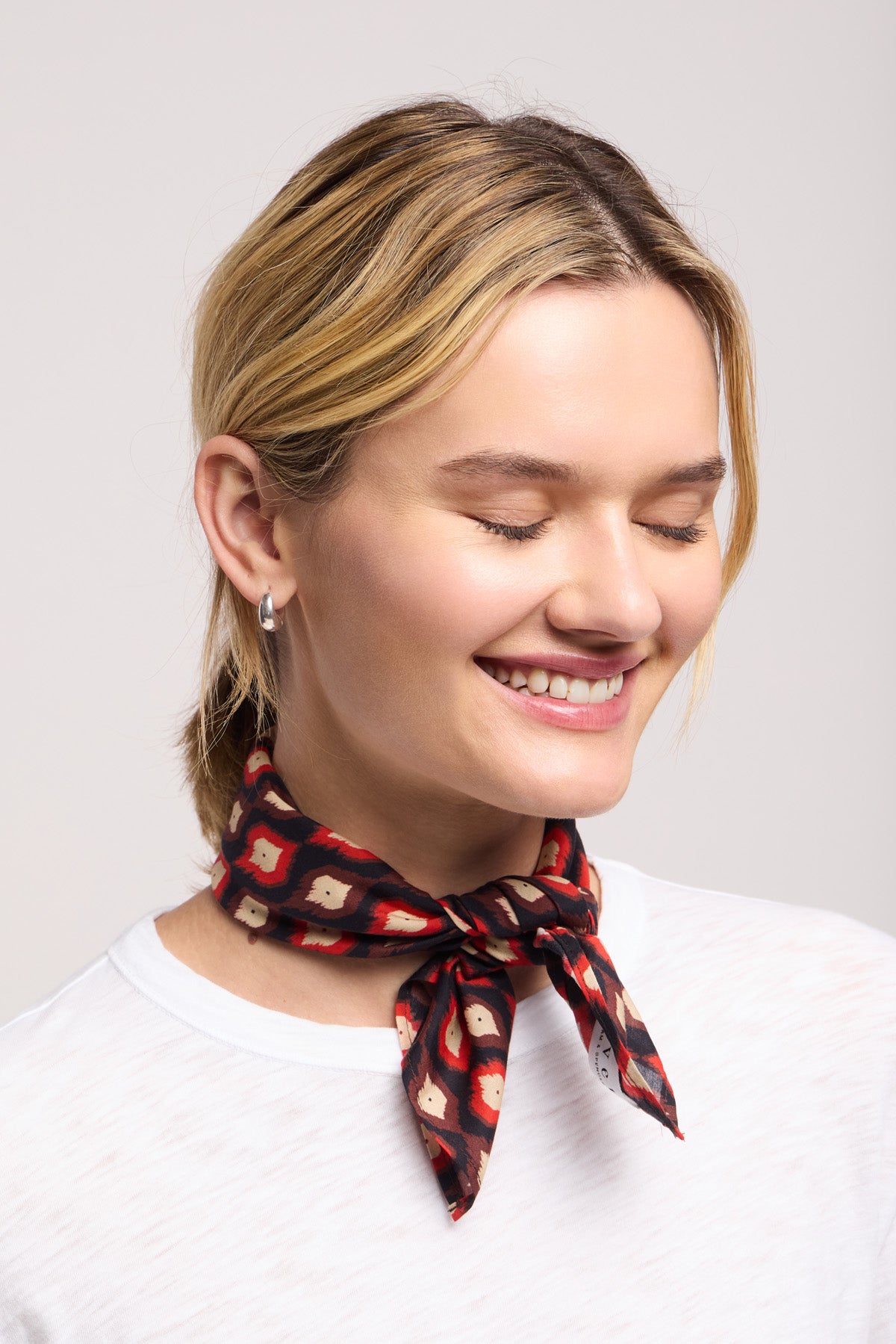 A woman with short blonde hair, in a white shirt and the JEWEL SCARF by Velvet by Graham & Spencer, smiles with her eyes closed against a plain background.