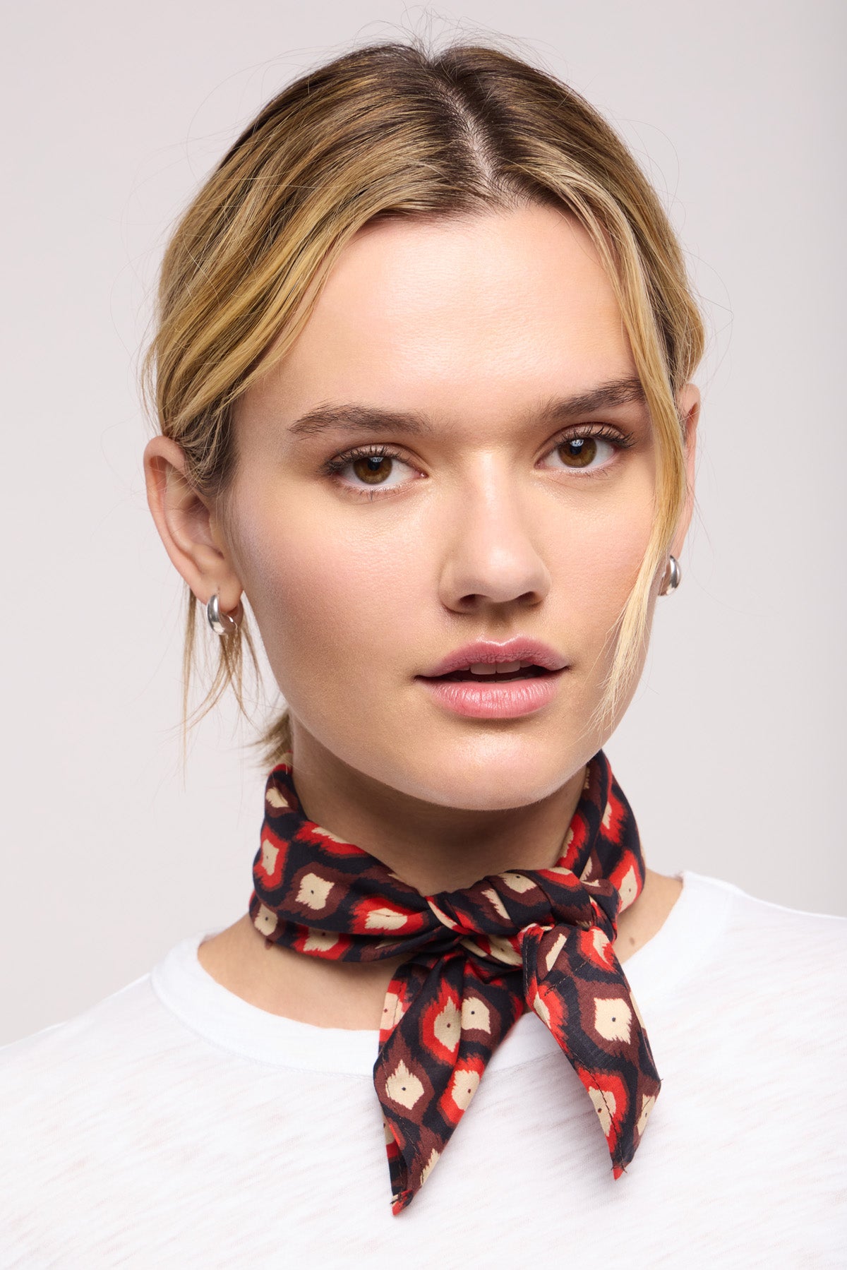 A person with light hair in a white shirt models the JEWEL SCARF by Velvet by Graham & Spencer, tied at the neck and looking at the camera against a plain background.-40453289935041