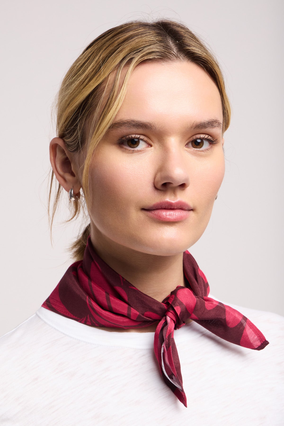A woman with light skin and blonde hair wears a white top and the JEWEL SCARF from Velvet by Graham & Spencer, featuring red patterns, tied around her neck as she looks directly at the camera against a plain background.-40453284167873
