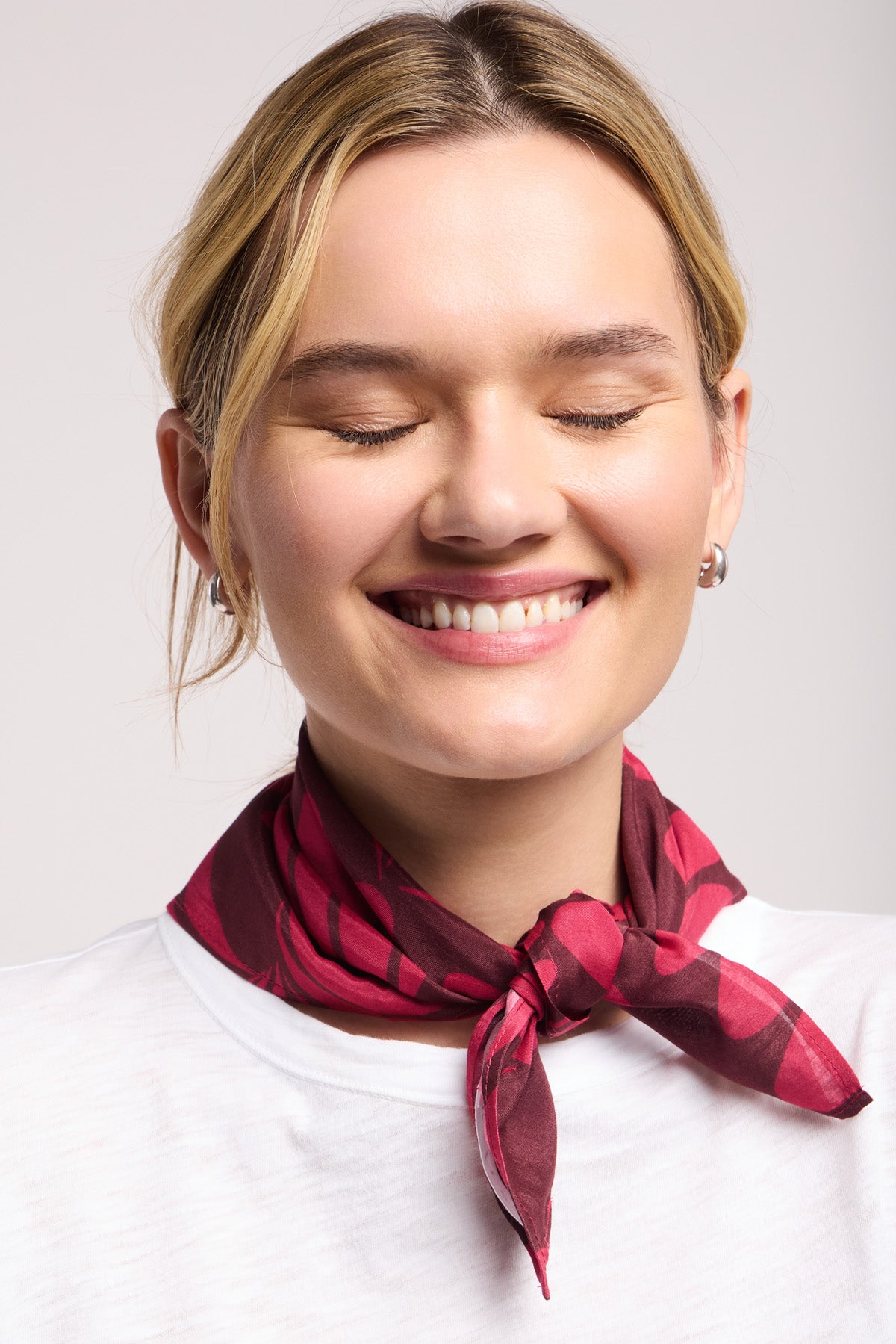 A woman with closed eyes and a smile wears the JEWEL SCARF by Velvet by Graham & Spencer—a red patterned accessory—paired with a white top against a plain background.-40453284102337