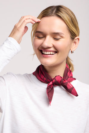 A woman with blonde hair smiles with her eyes closed, wearing a white top and the JEWEL SCARF by Velvet by Graham & Spencer—a chic red patterned accessory tied around her neck.