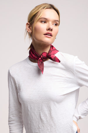 A person wearing a plain white long-sleeve shirt and the JEWEL SCARF by Velvet by Graham & Spencer—a chic red lightweight scarf tied around their neck—looks slightly to the side against a neutral background.