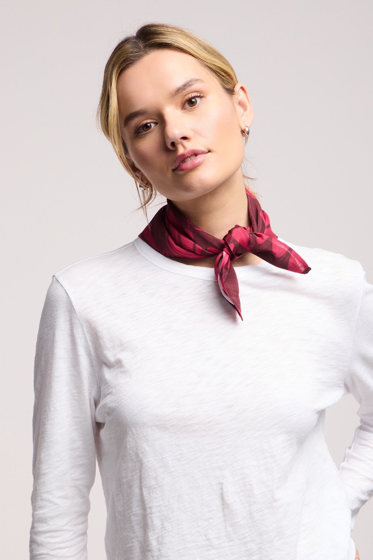 A person in a white long-sleeve shirt accessorizes with the Velvet by Graham & Spencer JEWEL SCARF—a red, lightweight scarf tied at the neck—standing against a plain light background.-40453284004033